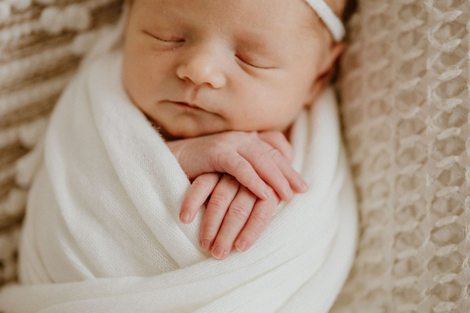 Closeup of sleeping newborn wrapped in white with hands crossed under chin on textured blanket.