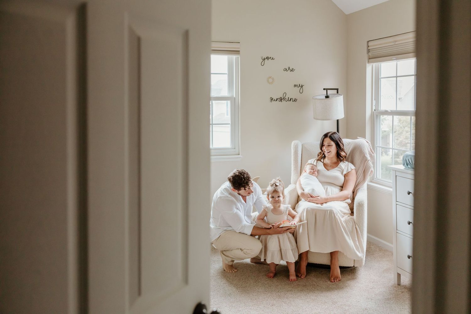 Intimate family moment captured in sunlit nursery room with white furniture.