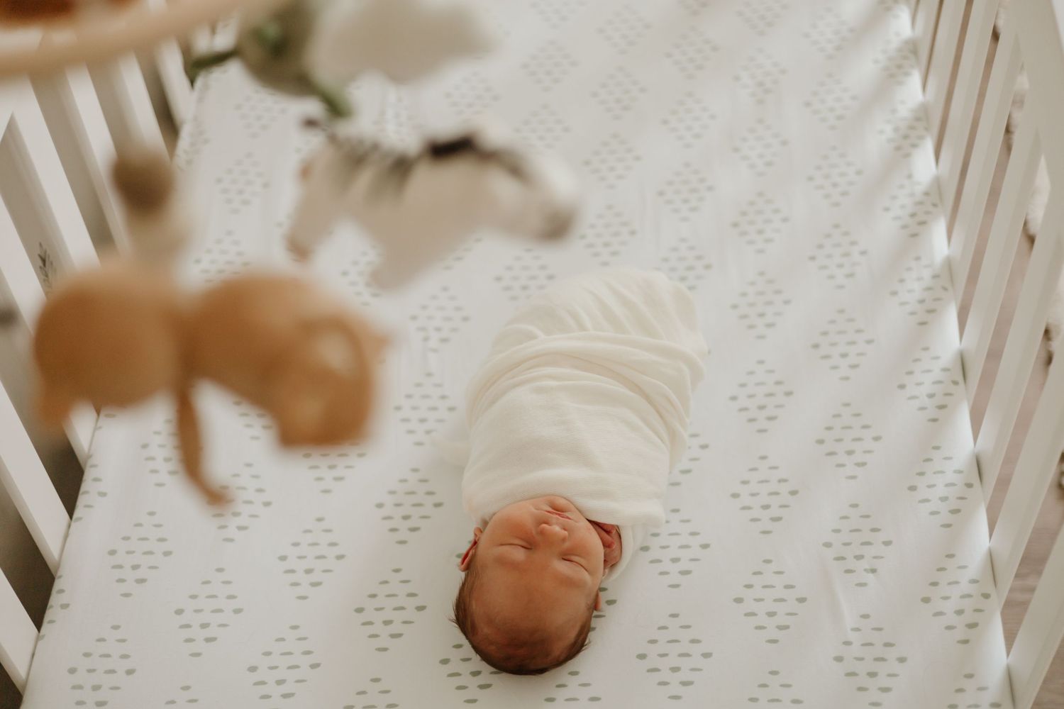 Overhead view of sleeping newborn in white crib with delicate diamond pattern bedding.