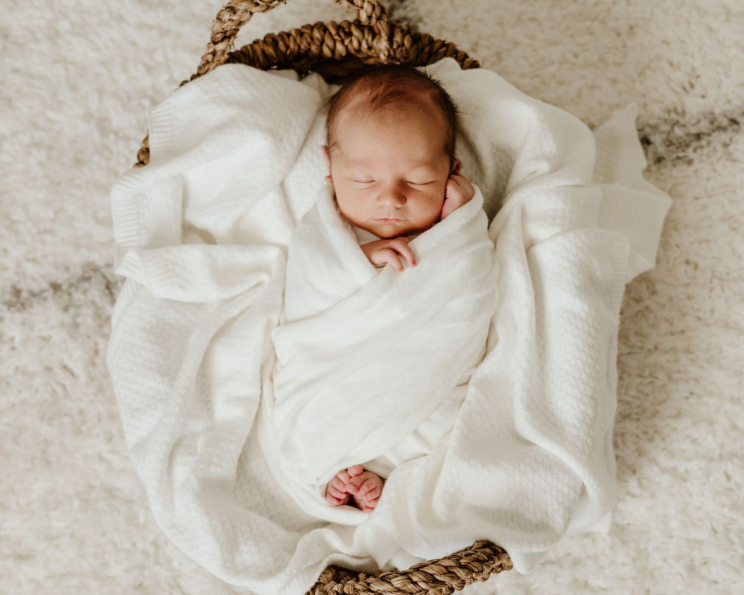 A peaceful sleeping newborn wrapped in white blanket rests in woven basket on carpet.