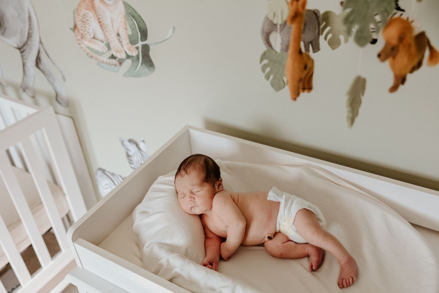 Sleeping newborn rests peacefully in white crib under jungle animal mobile.