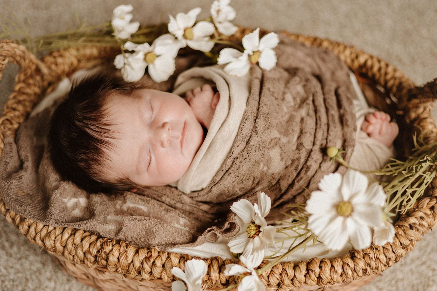 A sleeping baby wrapped in brown swaddle rests in woven basket surrounded by white flowers.