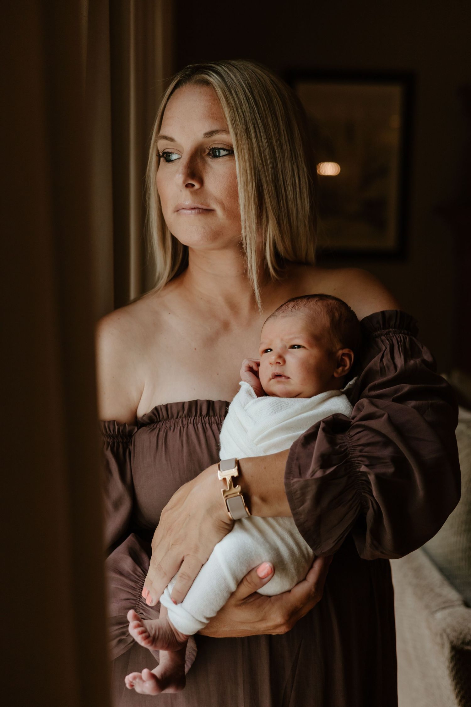 Mother in brown off-shoulder dress holding sleeping newborn baby wrapped in white.