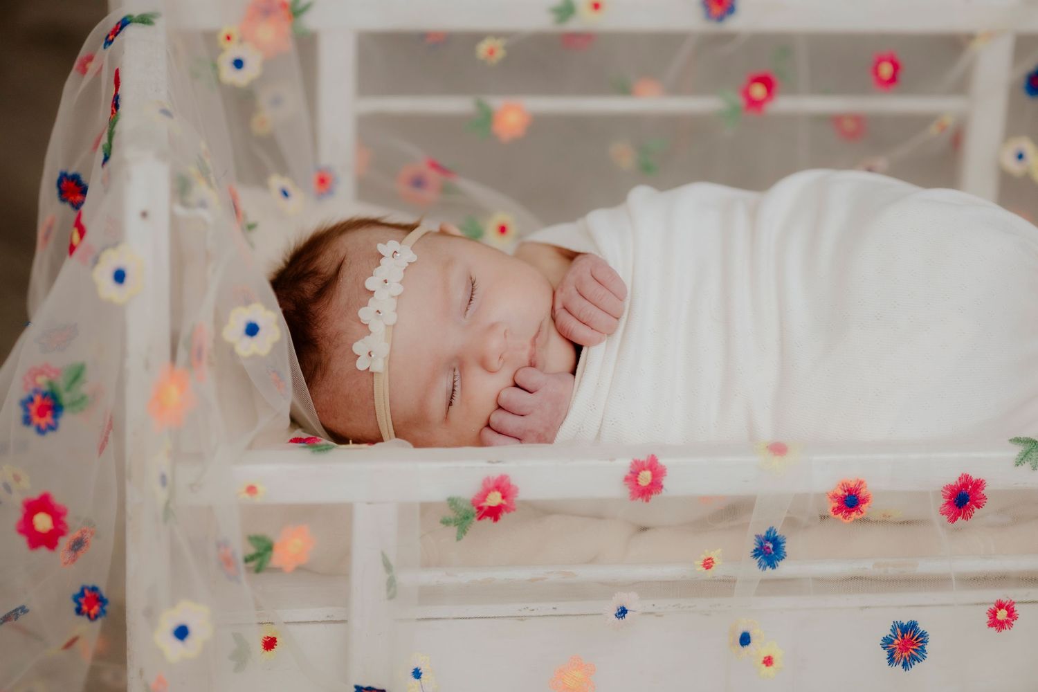 Sleeping newborn in white wrap surrounded by colorful floral lights and decorations.