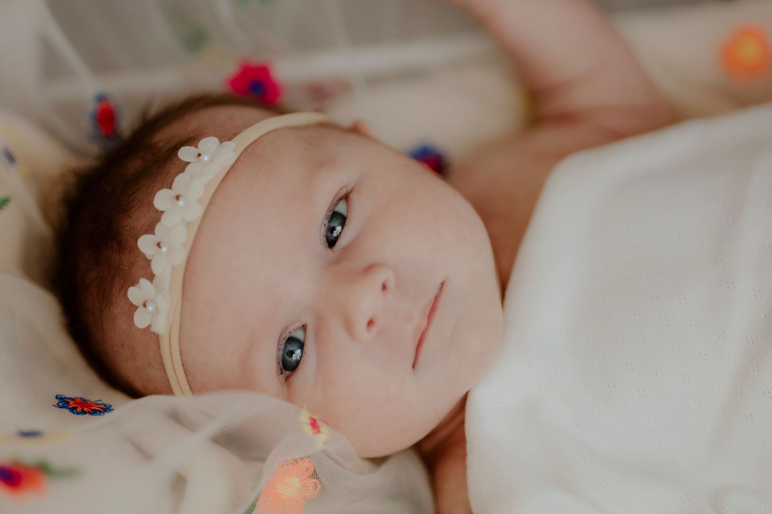 Baby wearing lace headband lying on white bedding with colorful string lights.