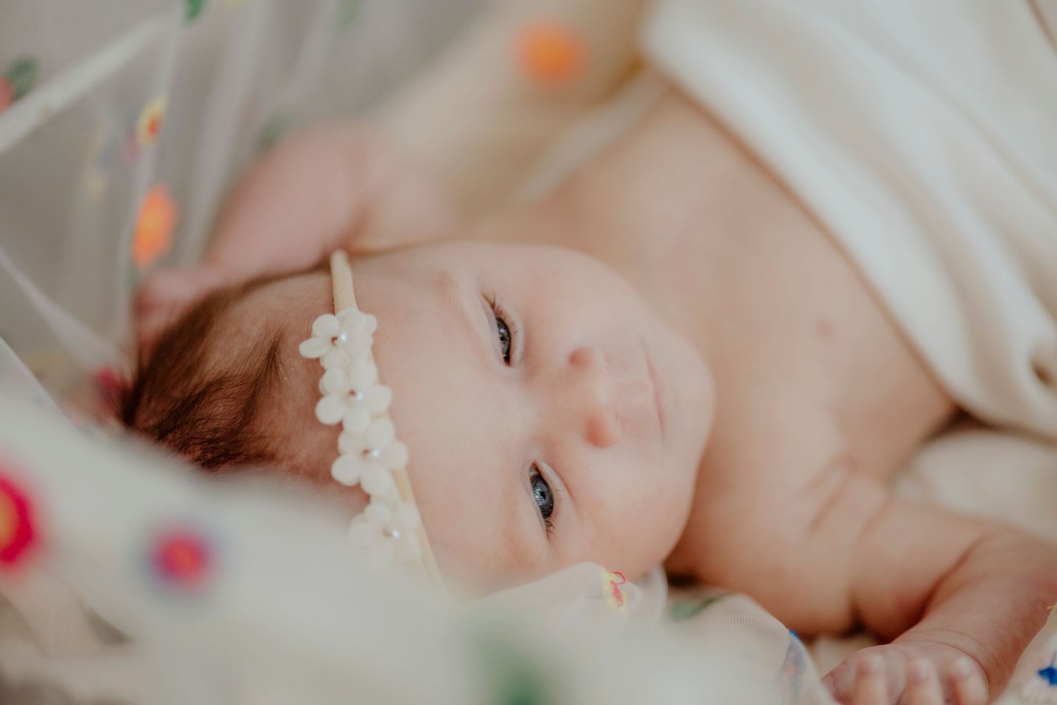 Baby in white headband resting on soft bedding with colorful floral lights.