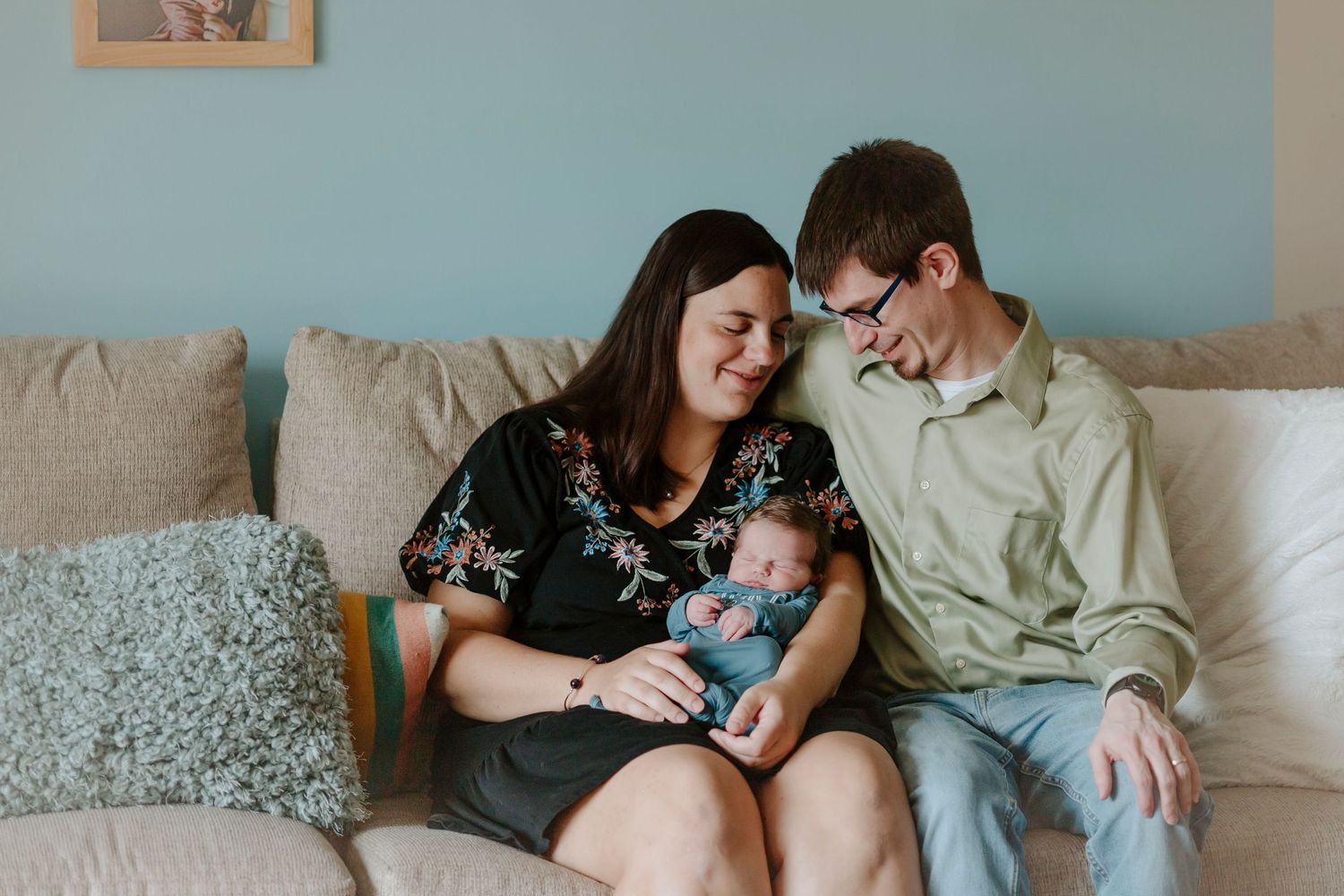 A couple sits on a gray couch sharing a tender moment with their newborn baby in a casual home setting.
