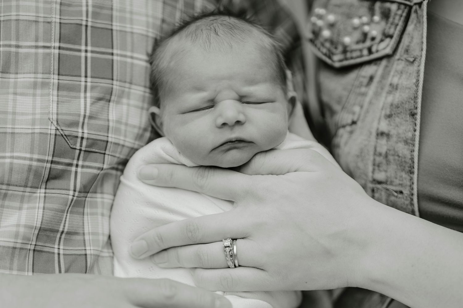 Black and white image of newborn baby swaddled in blanket with hand wearing ring visible.