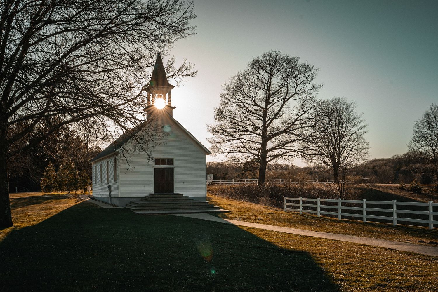 Small white historic church glowing in golden sunset light surrounded by bare trees.
