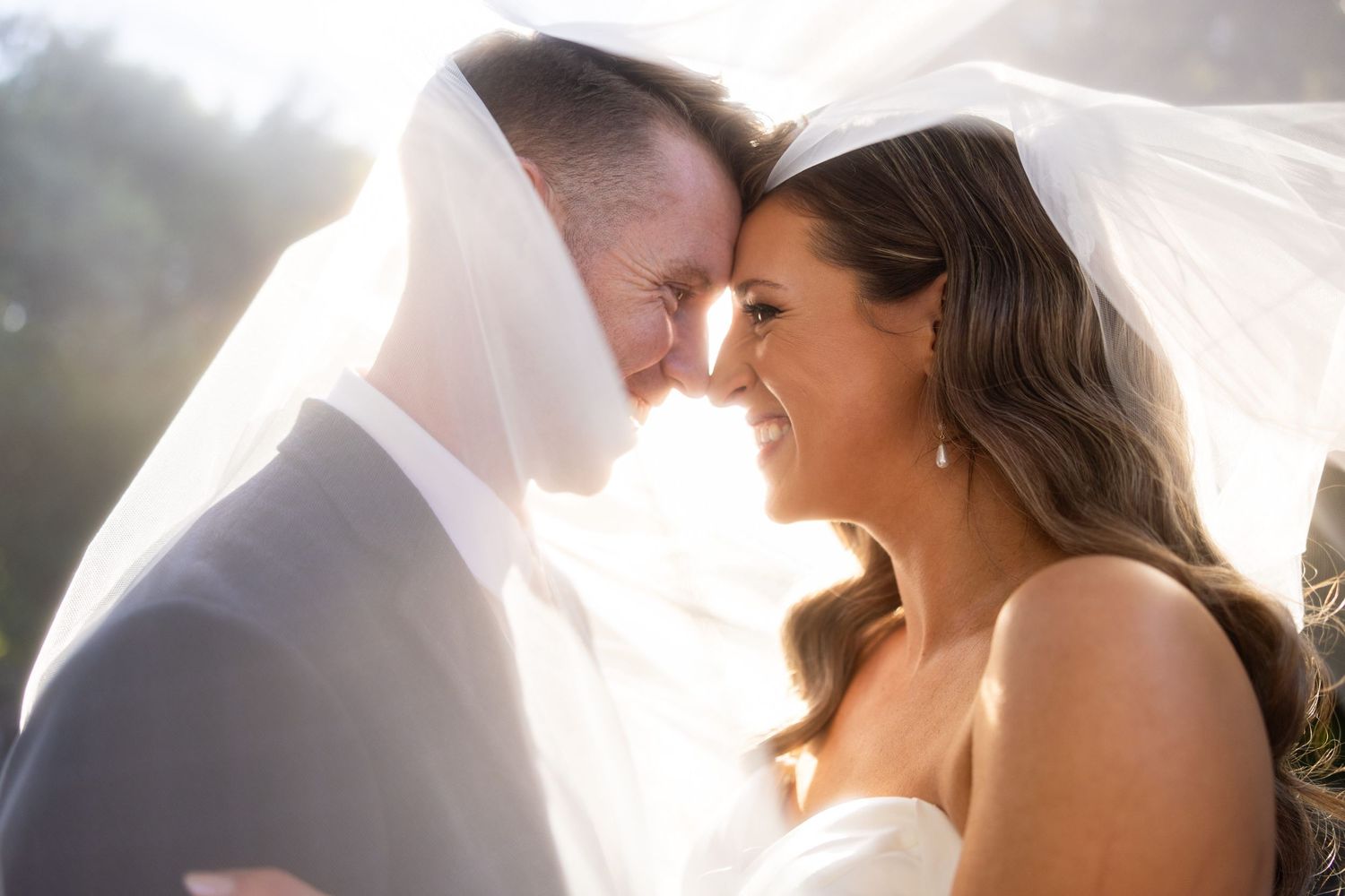 Romantic wedding moment with couple sharing an intimate look under a flowing veil in sunlight.