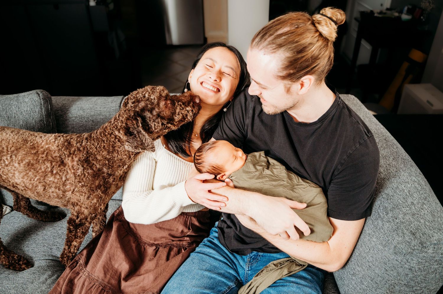 Mississauga newborn photographer captures new parents holding baby on couch while dog joins in to give kisses