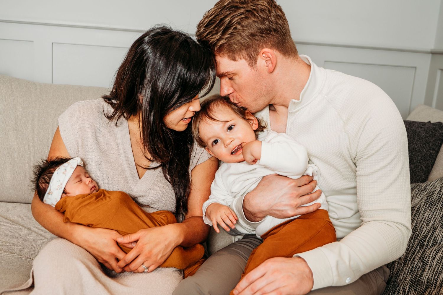 Mom and dad sit together on couch in their home while holding toddler and newborn baby.