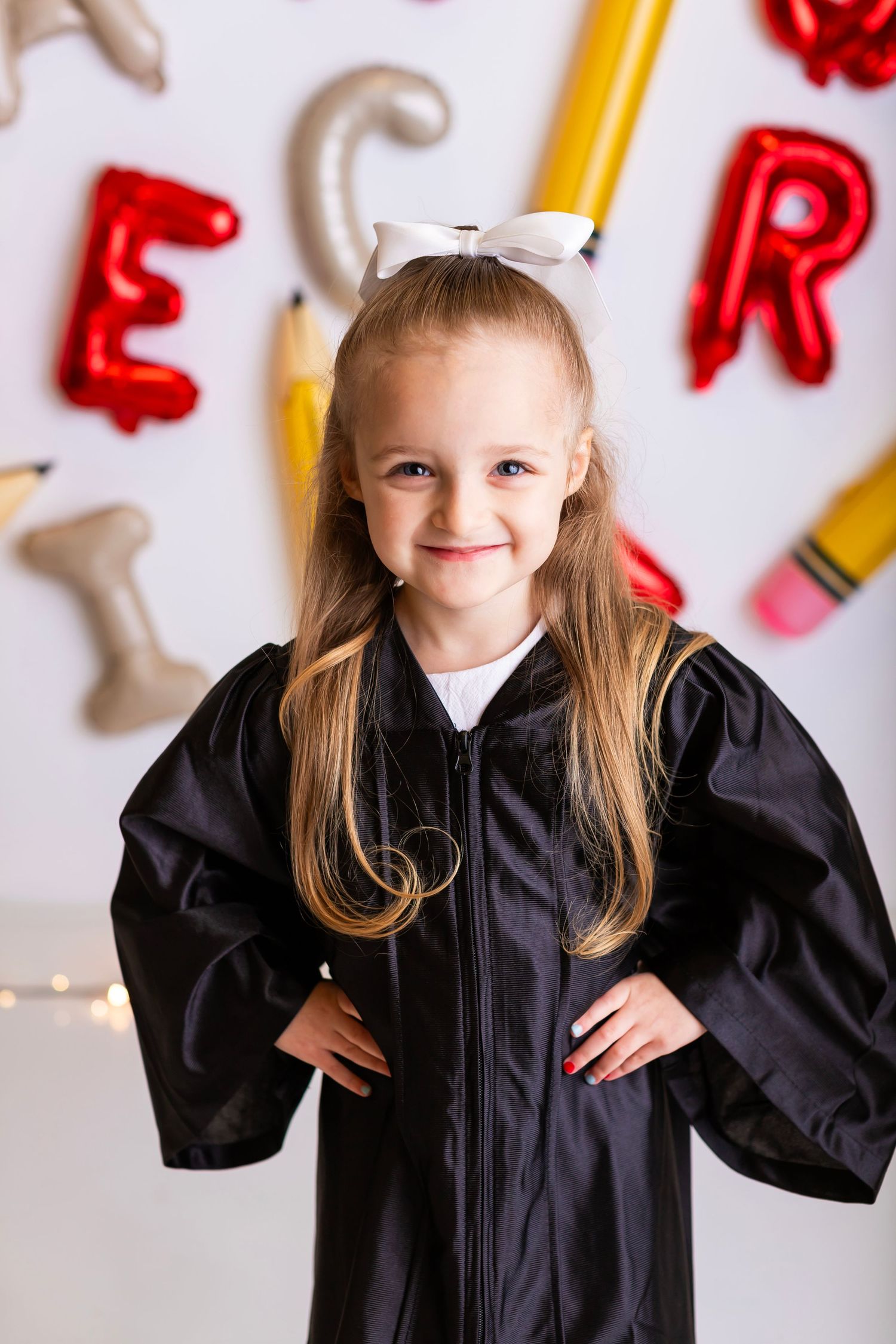 Young graduate in black cap and gown posing against wall with decorative letters and pencils.