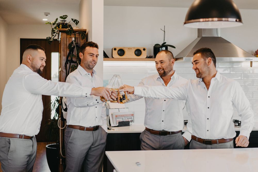 Men in white shirts and grey pants toasting with drinks in modern kitchen.