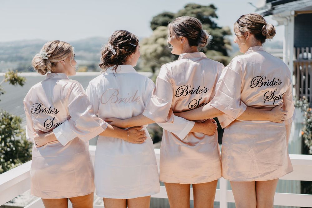Four women wearing matching white robes with 'Bride's Crew' text standing on balcony.