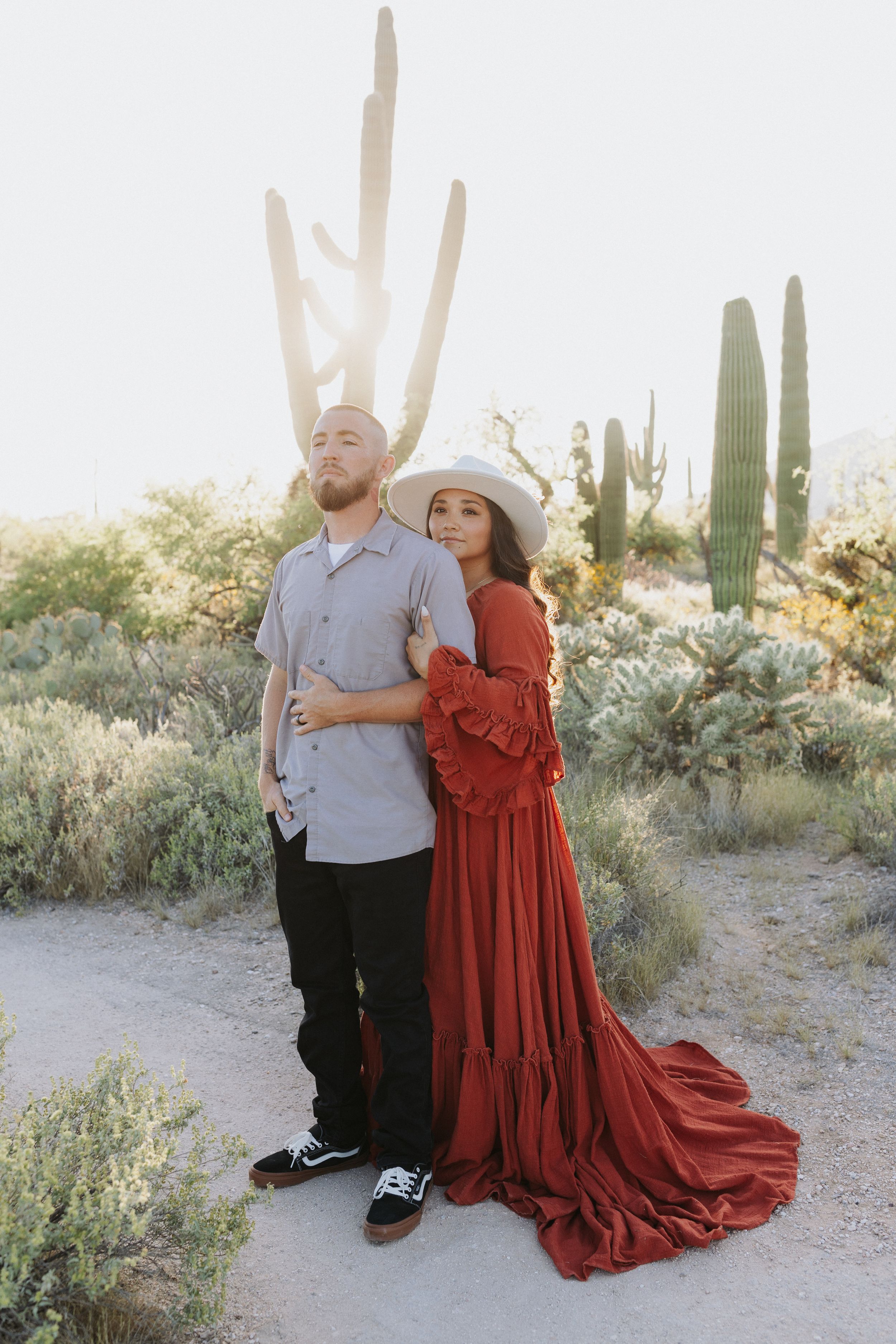 A couple poses in front of saguaro cacti at sunset, with her wearing a dramatic red dress.