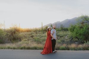 A couple stands together on a desert road with mountains and cacti in the background.