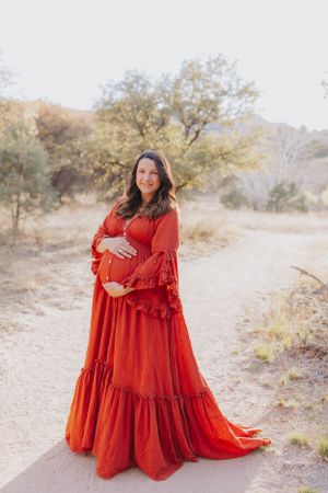 Expectant mother in flowing red dress poses on desert path with mountains behind.