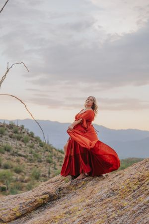 A red dress flows dramatically as someone poses on a rocky desert slope at dusk.