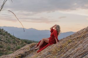Someone in a red dress reclines on a rocky desert slope during sunset.