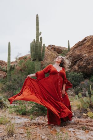 A red dress billows in the wind among towering saguaro cacti in a desert setting.