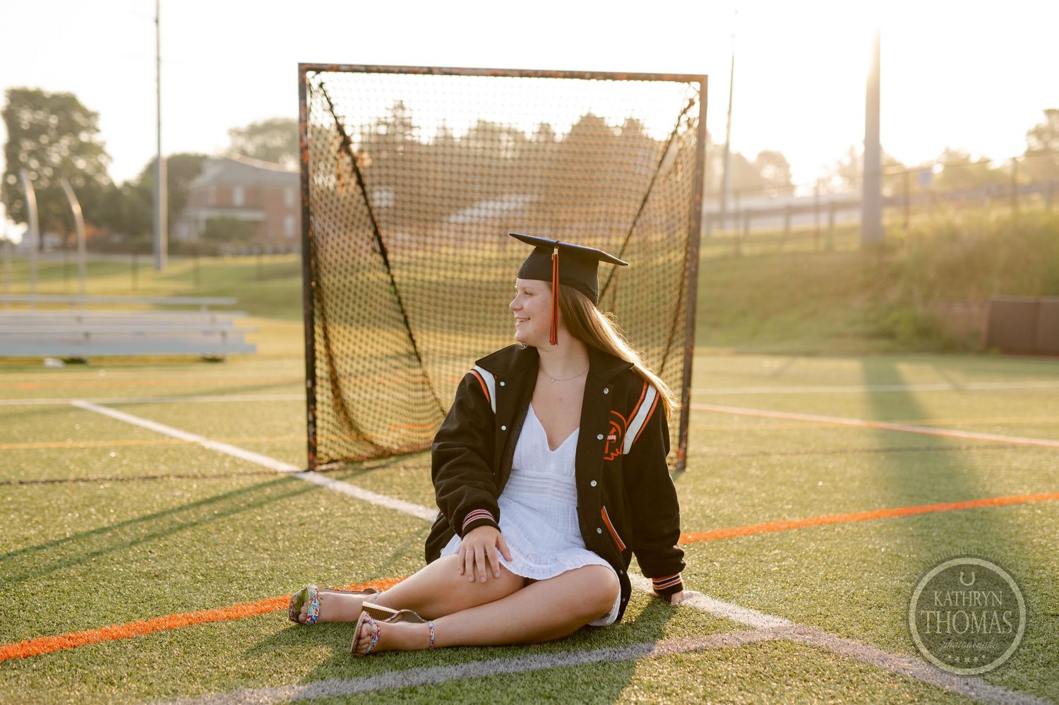 Graduate in cap and gown sits by lacrosse goal during golden hour photo session.