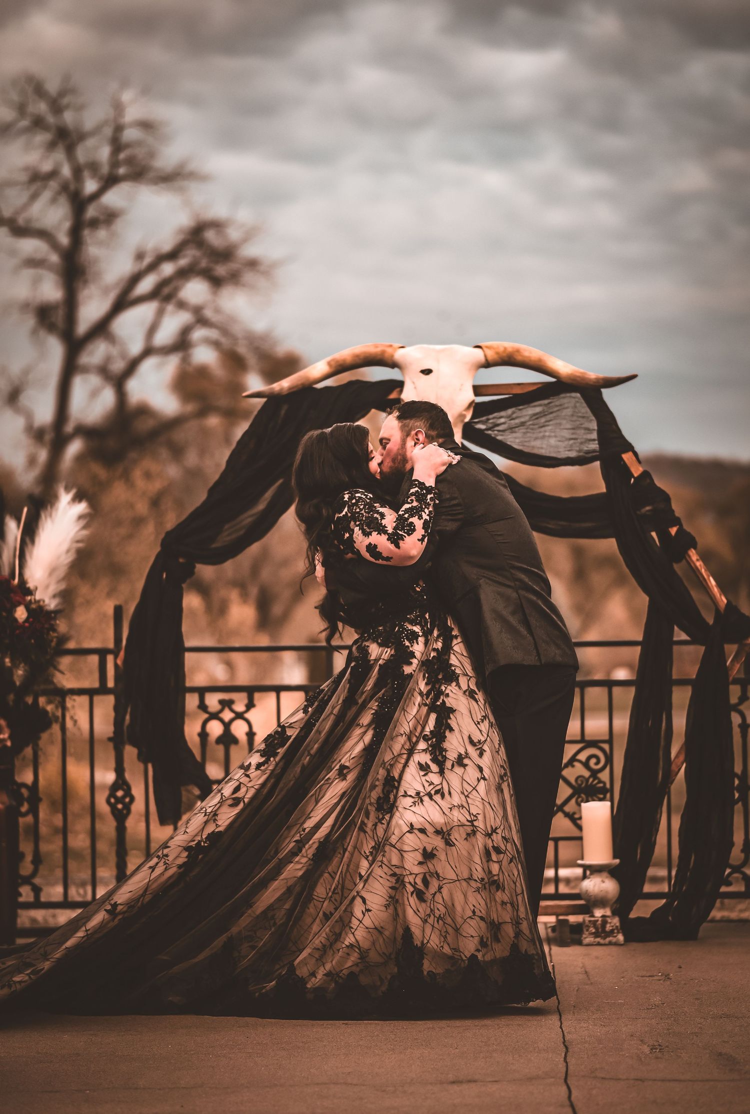 A dramatic wedding photo showing a couple in black attire embracing under a towering animal skull arch.