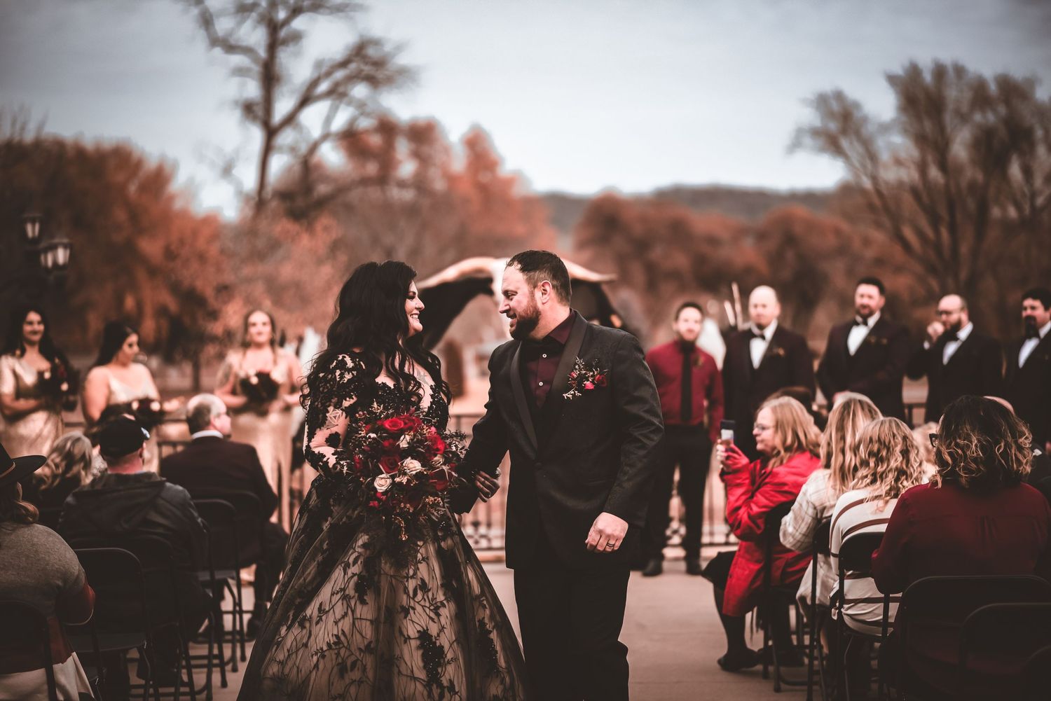 A gothic wedding ceremony with guests watching the couple kiss in front of autumn trees.