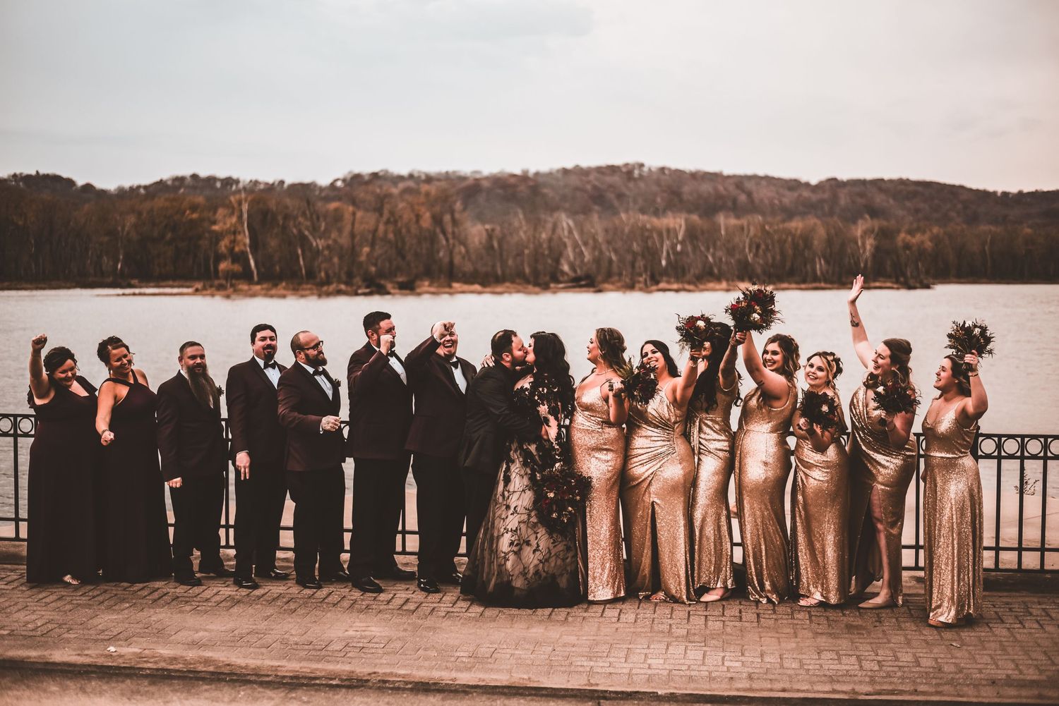 Wedding party poses together on waterfront deck with autumn foliage in background.