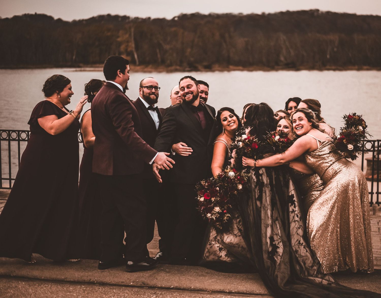Wedding party shares candid moment of laughter on lakeside deck at sunset.
