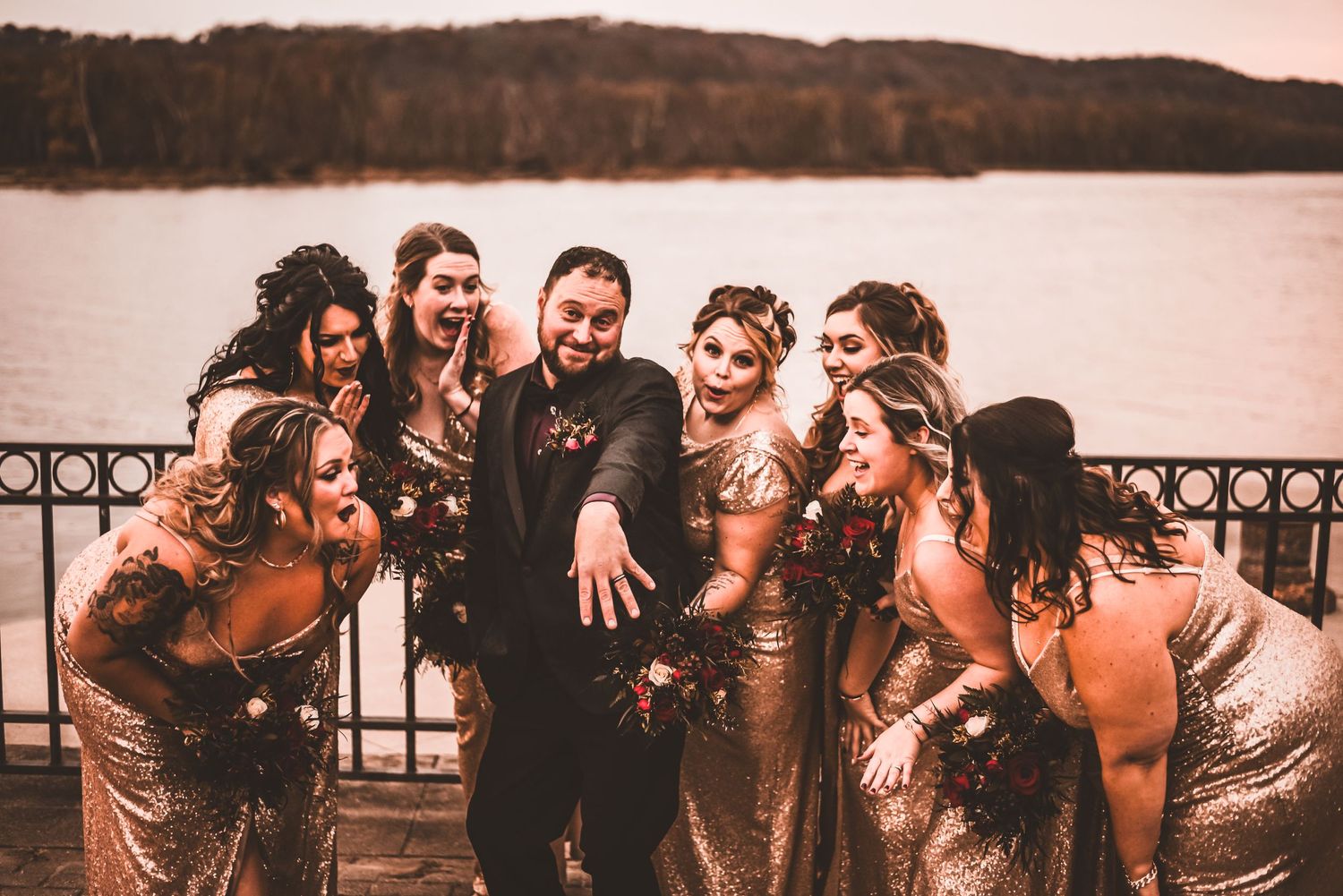Wedding party in sparkly gold dresses poses by lakeside railing at sunset.