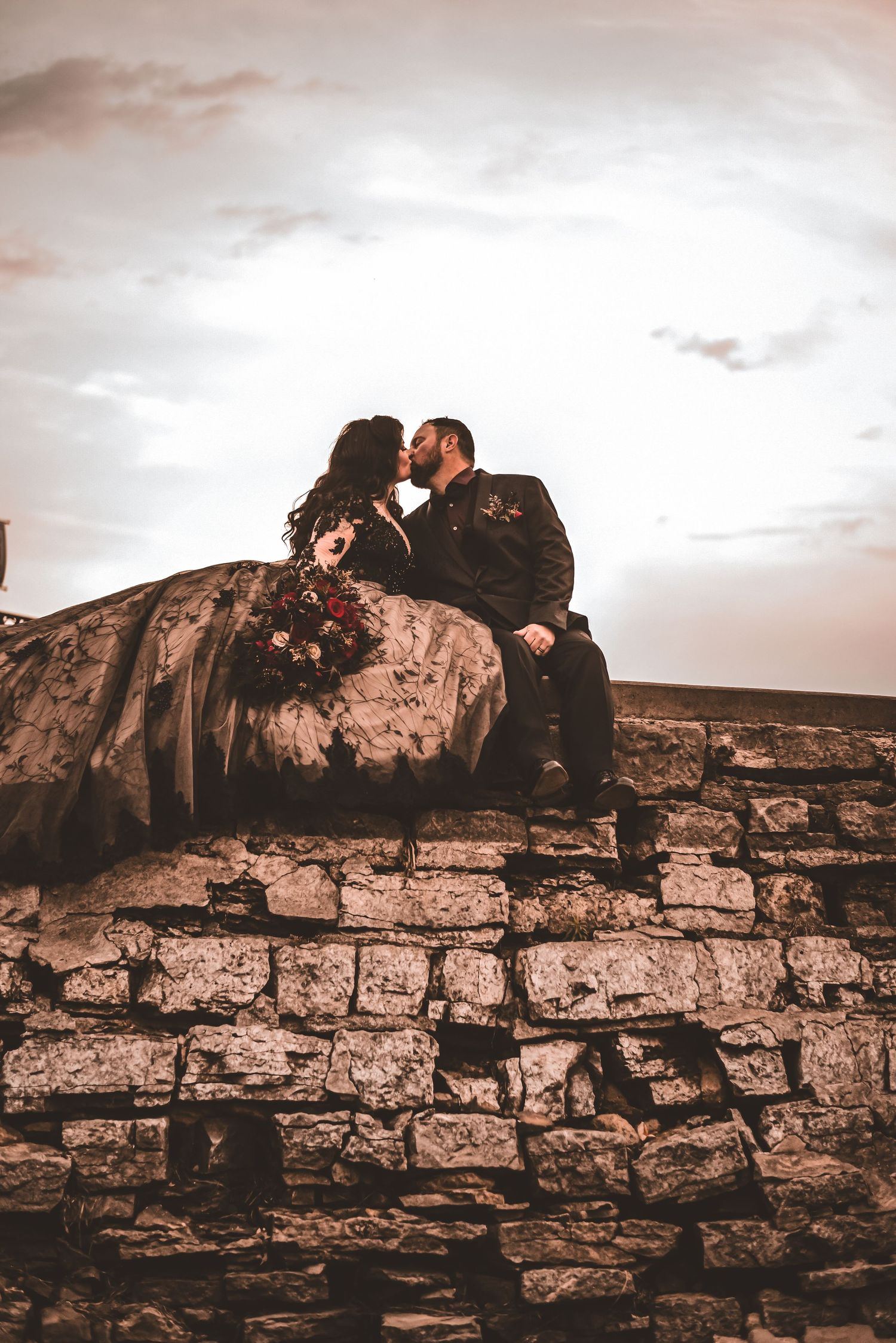 A romantic sunset portrait of newlyweds sitting on stone wall in gothic wedding attire.