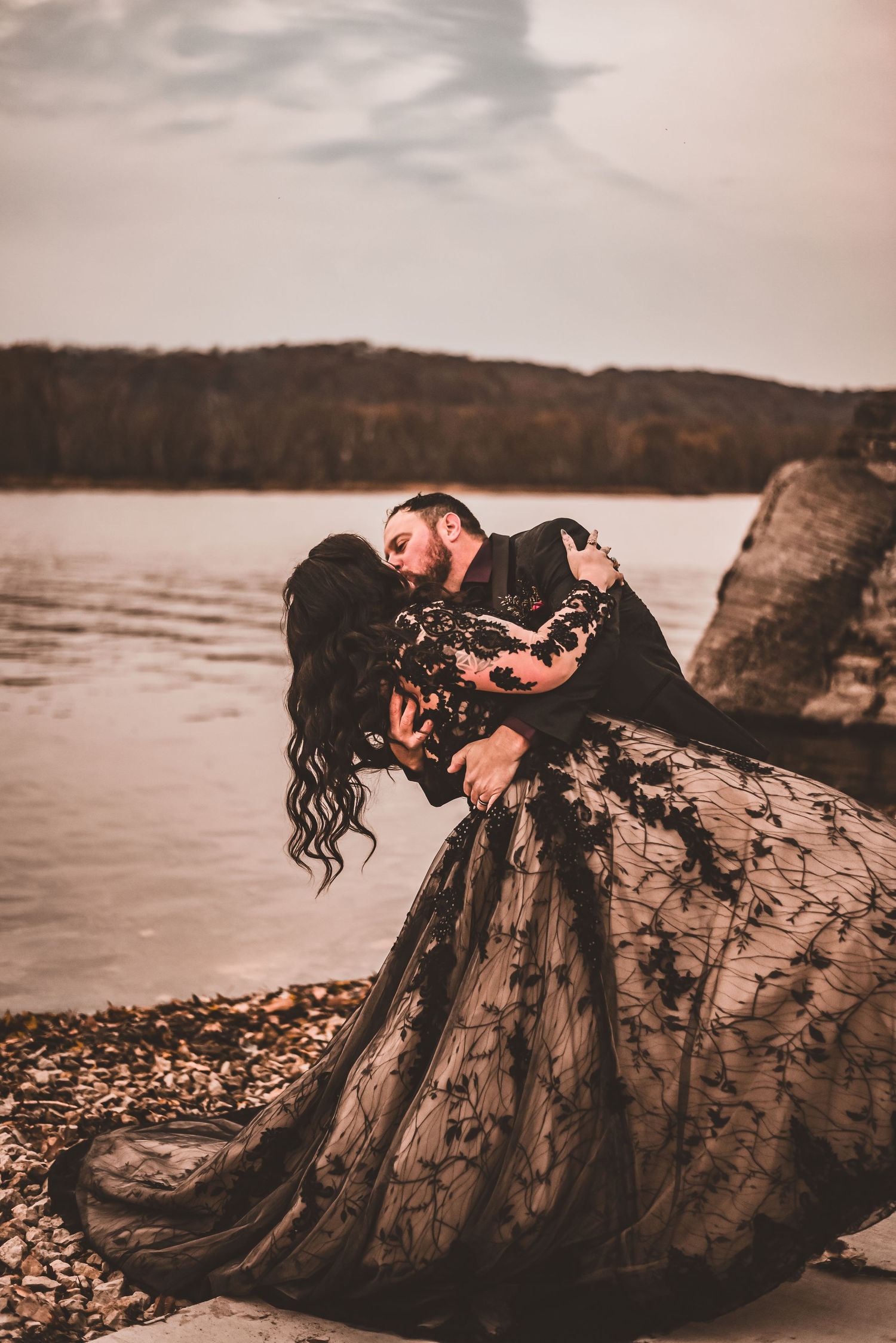 A dramatic dip pose by a couple in black wedding attire near a waterfront at sunset.