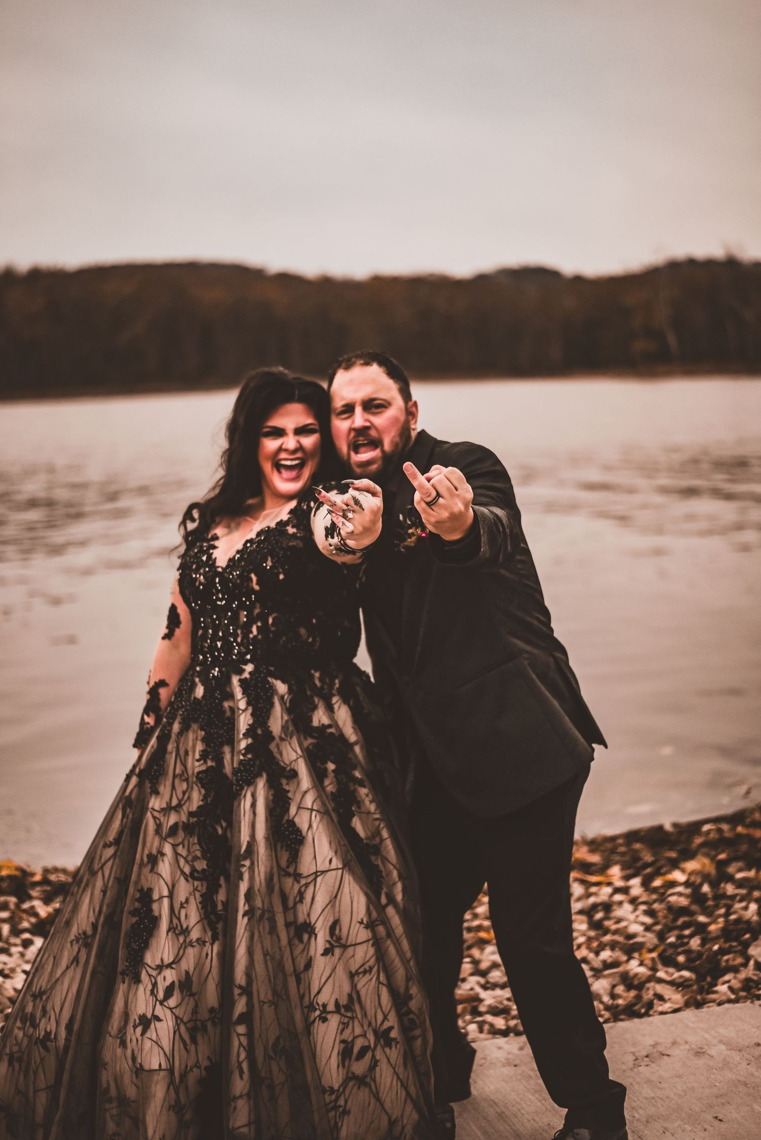 A couple in formal attire poses playfully by a lake, with the woman wearing a black lace gown.