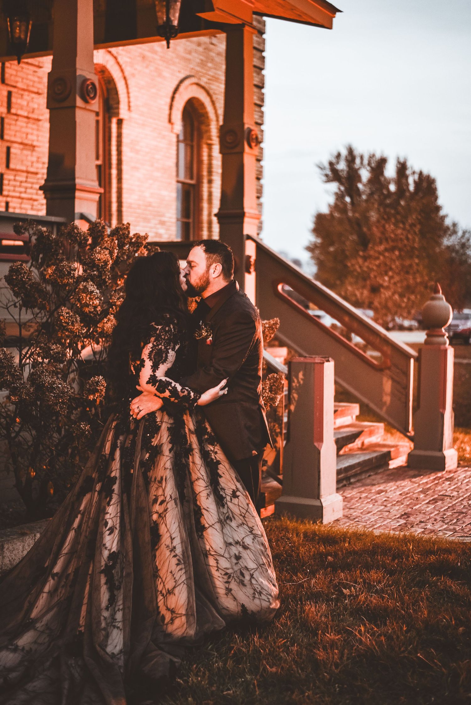 A couple embraces in formal wear outside a historic brick building during sunset.
