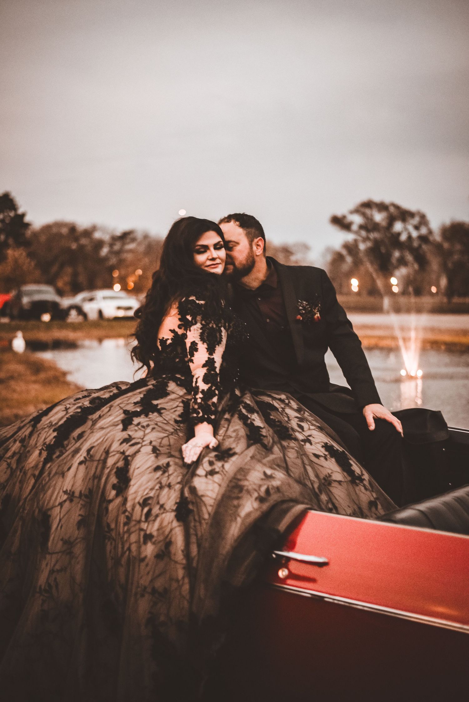 A romantic couple poses by a fountain at sunset, with the woman wearing an elegant black lace ball gown.