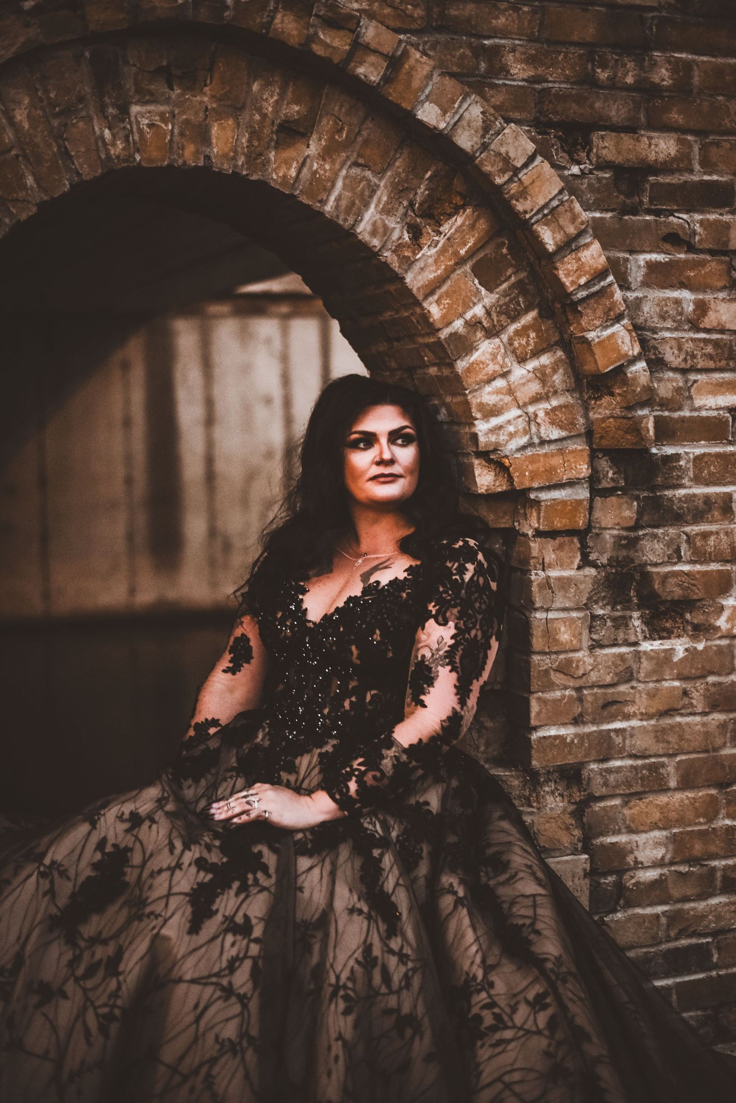 A woman in a black lace ball gown poses under a rustic brick archway.