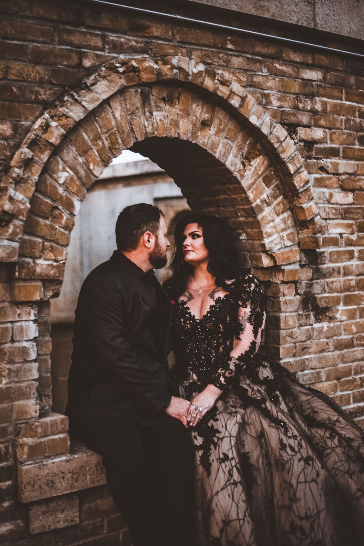 A romantic couple embraces under a vintage brick archway, with the woman wearing a dramatic black gown.