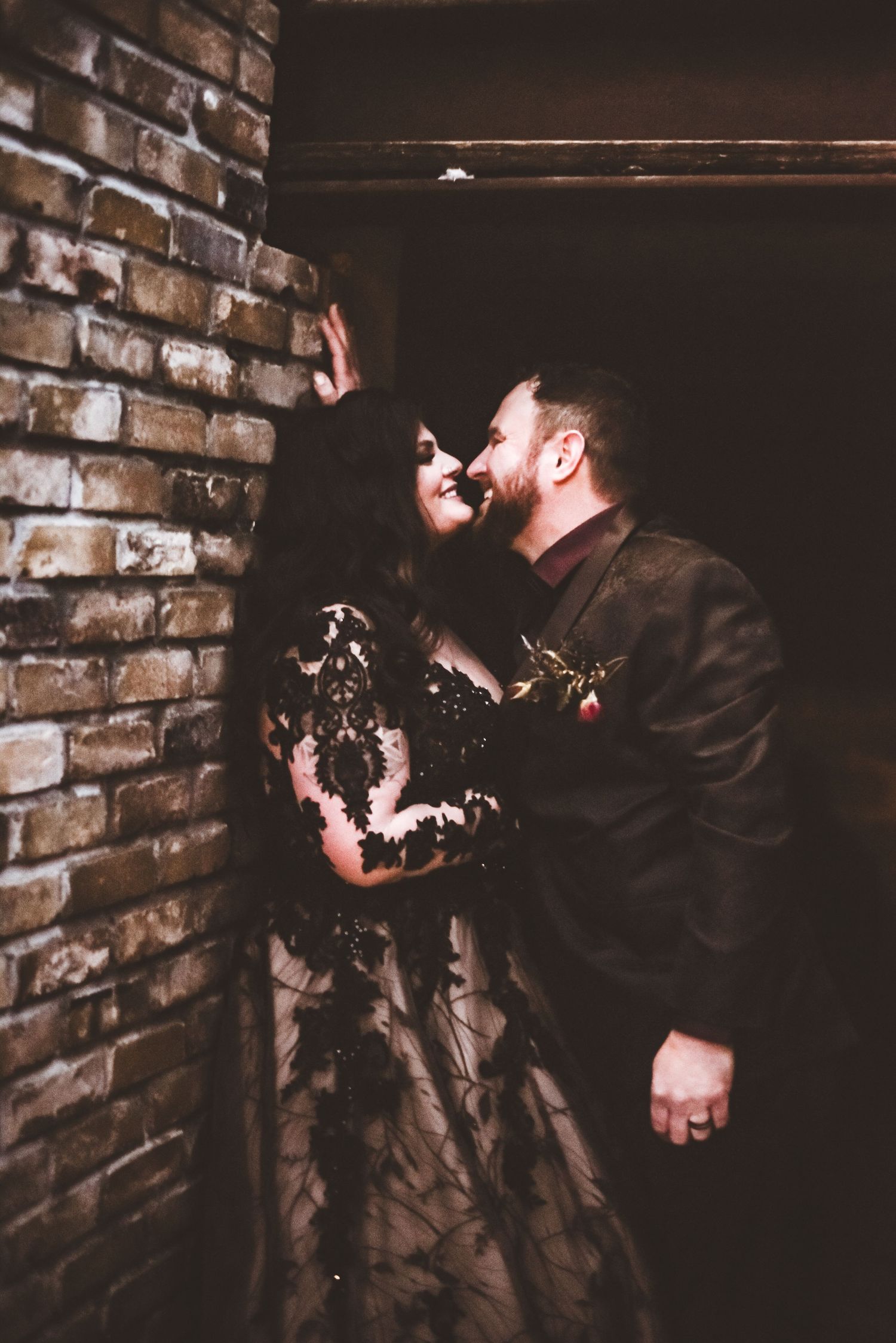 A couple shares an intimate moment against exposed brick walls in vintage-inspired formal attire.
