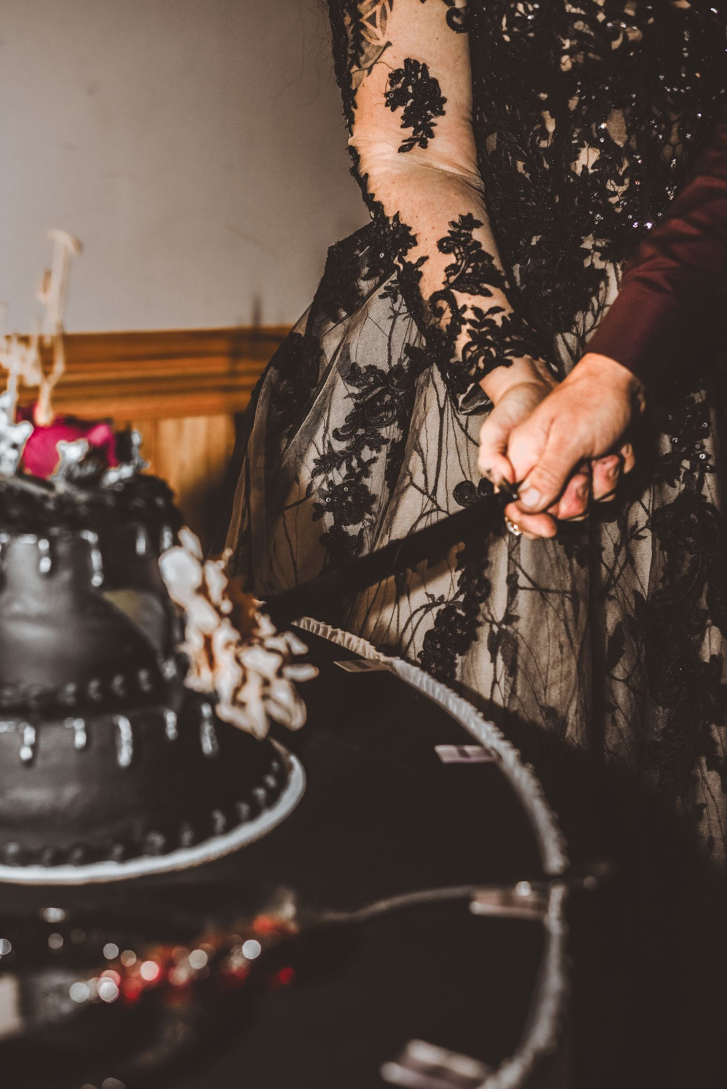 Close-up of black lace wedding dress details next to a dark tiered cake.