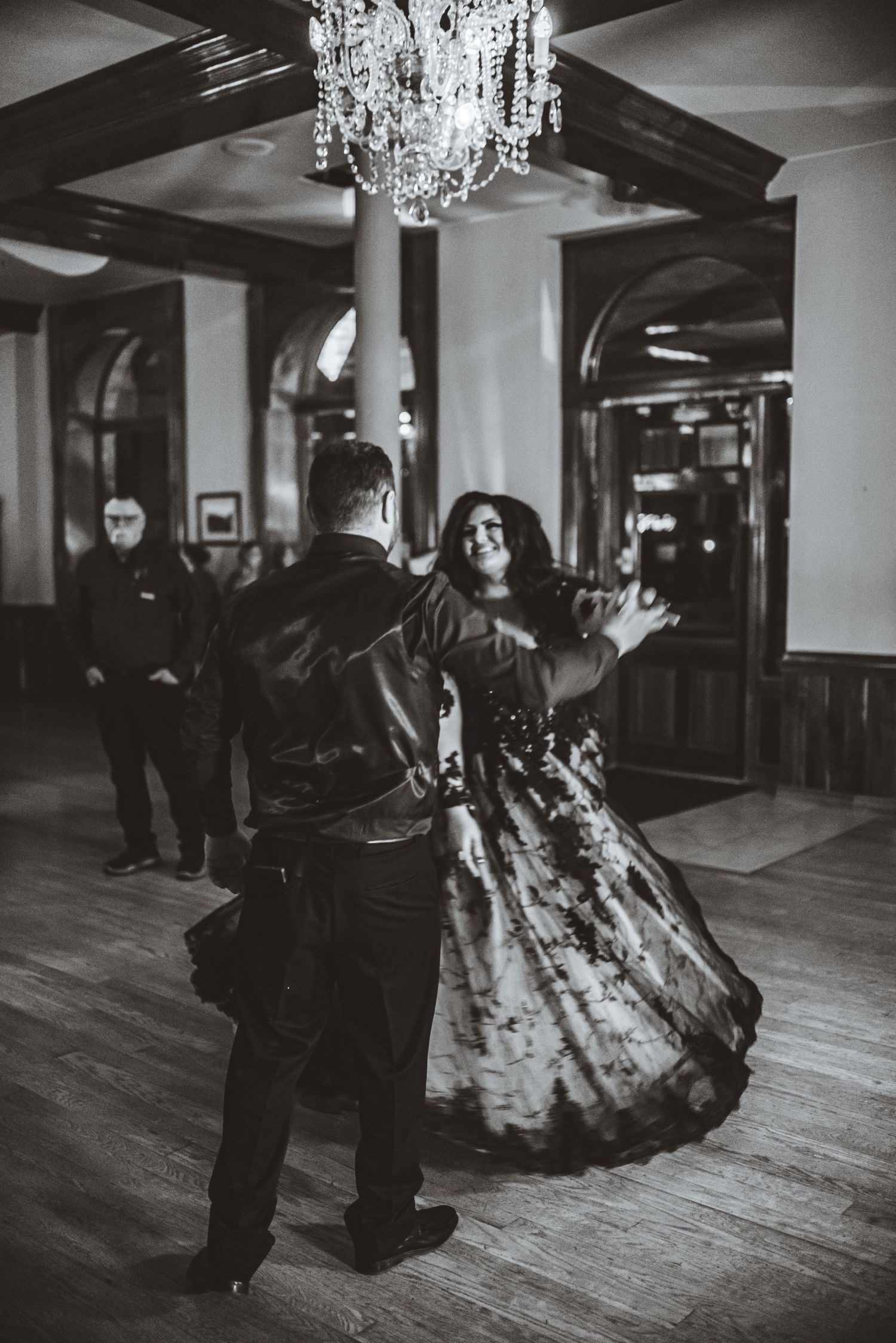 A dancing couple twirls under a crystal chandelier in a classic ballroom setting.