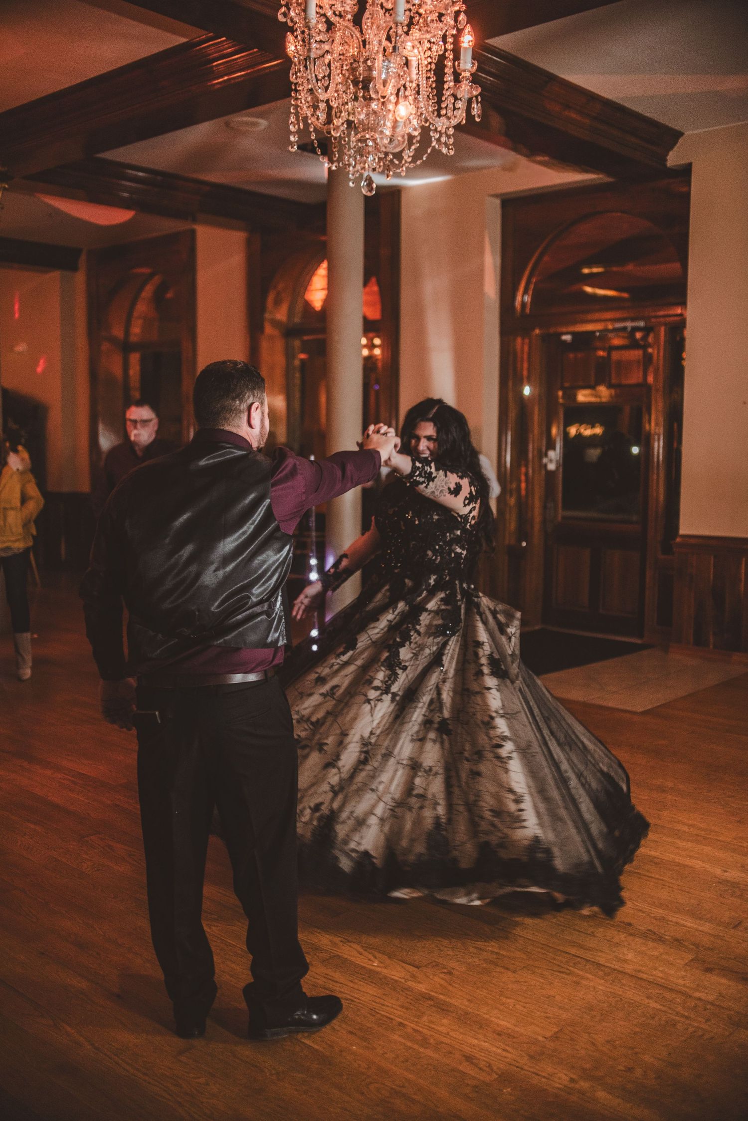 A couple shares a dance under ornate lighting in an elegant venue with wooden floors.