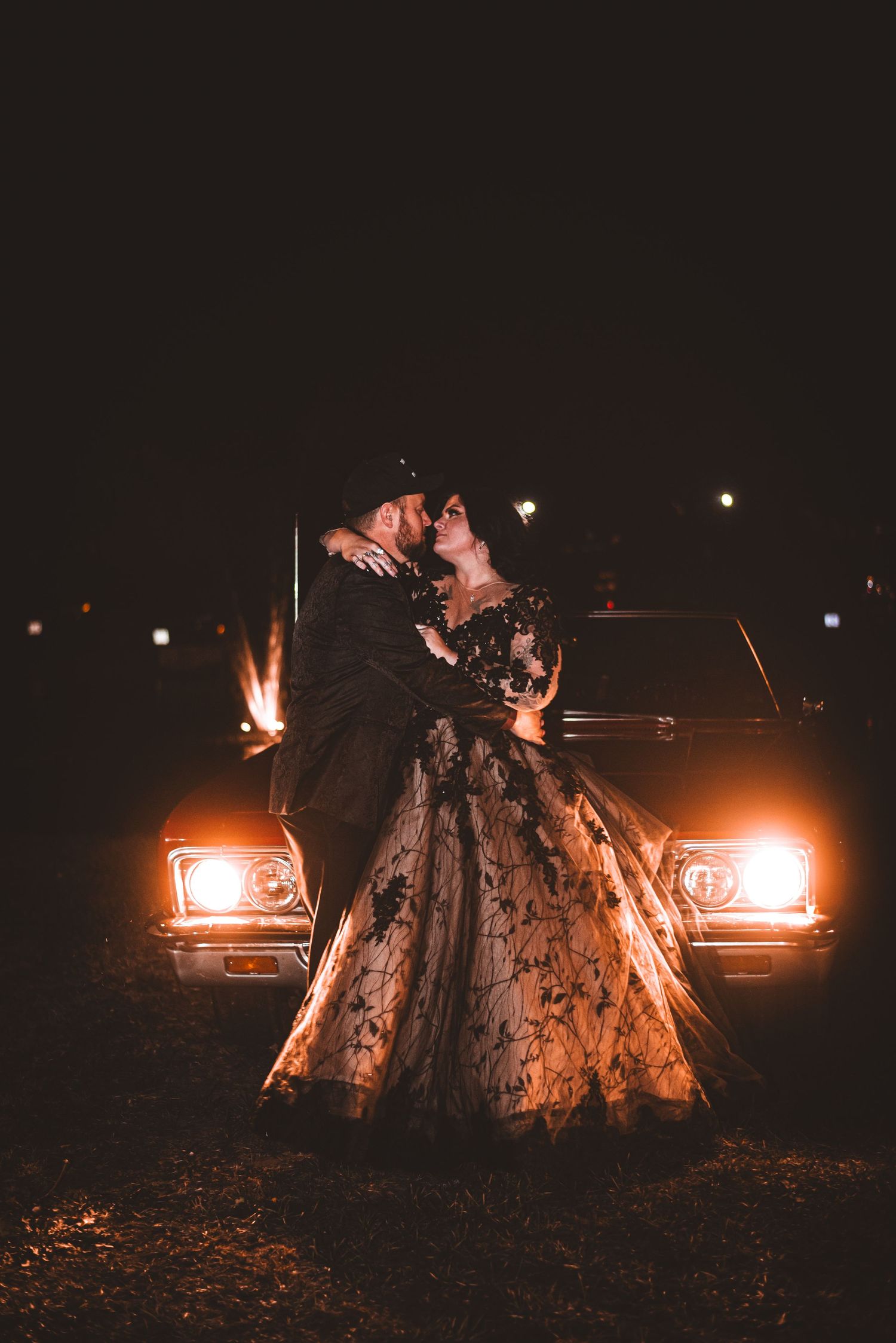 Romantic couple poses in front of vintage car headlights at night with a dramatic formal dress.