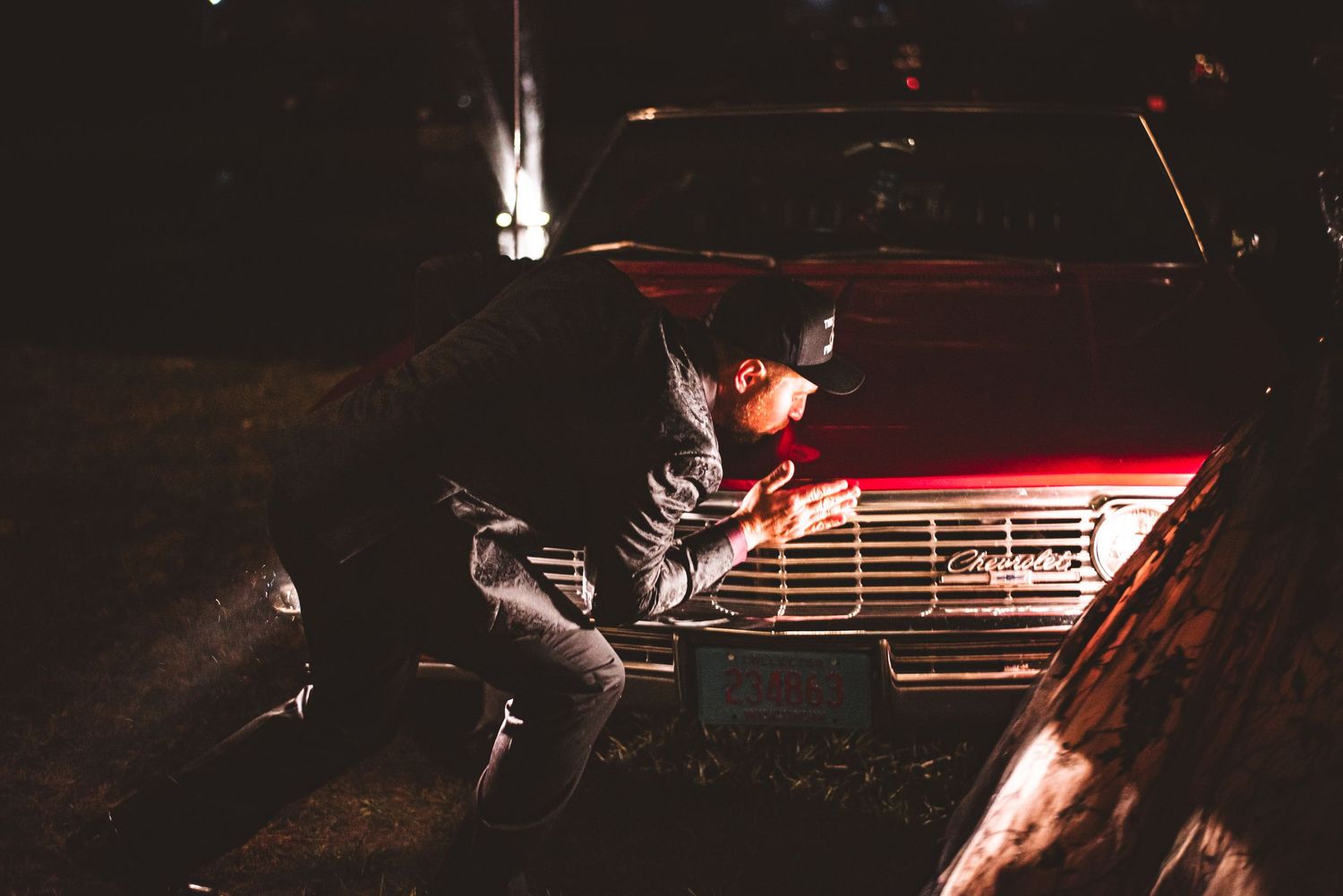 Person in dark clothing poses on vintage car hood with red lights in nighttime setting.