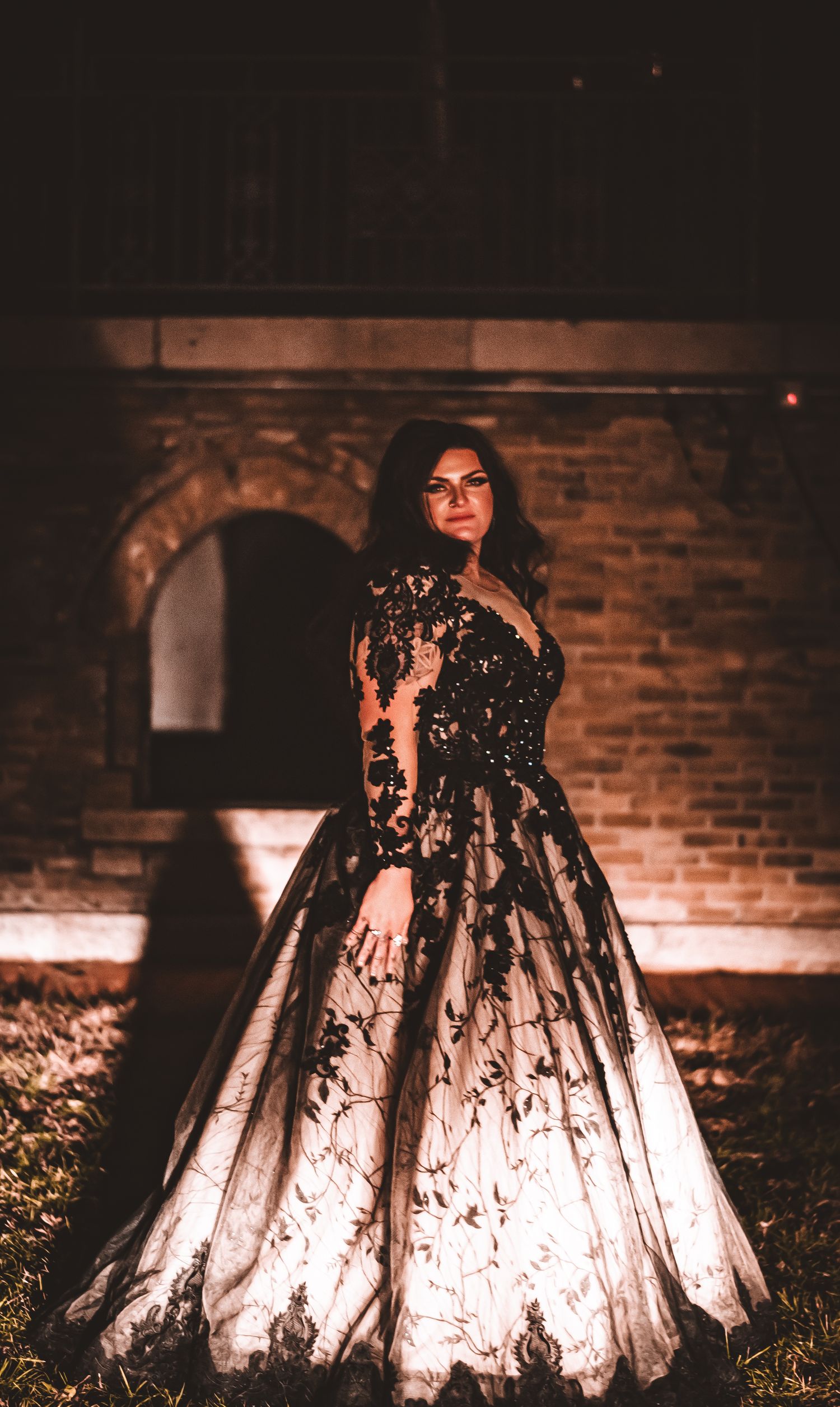 Woman in black lace ball gown poses against brick wall at night with dramatic lighting.