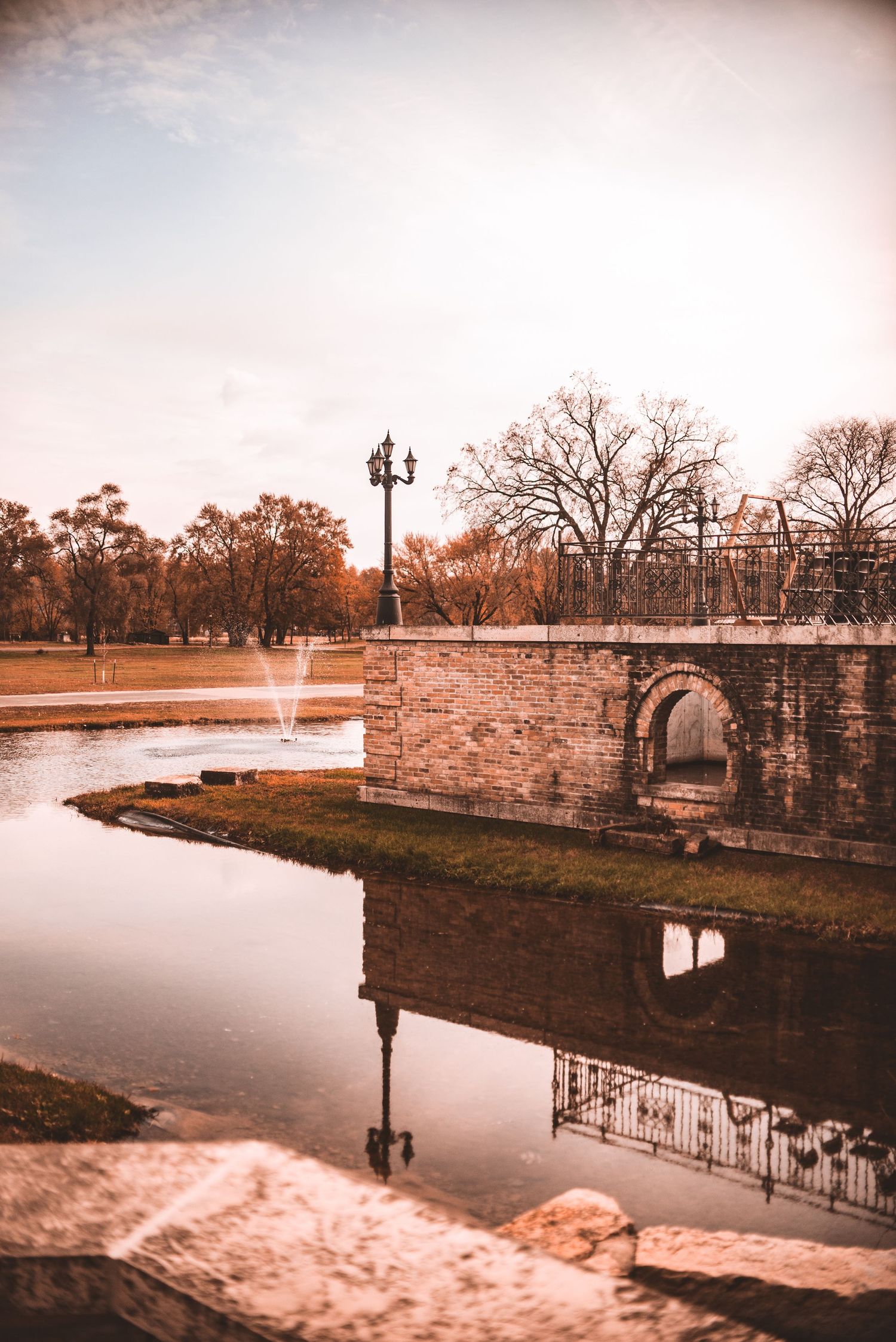 A serene landscape with a stone bridge, fountain, and reflection in water during golden hour.