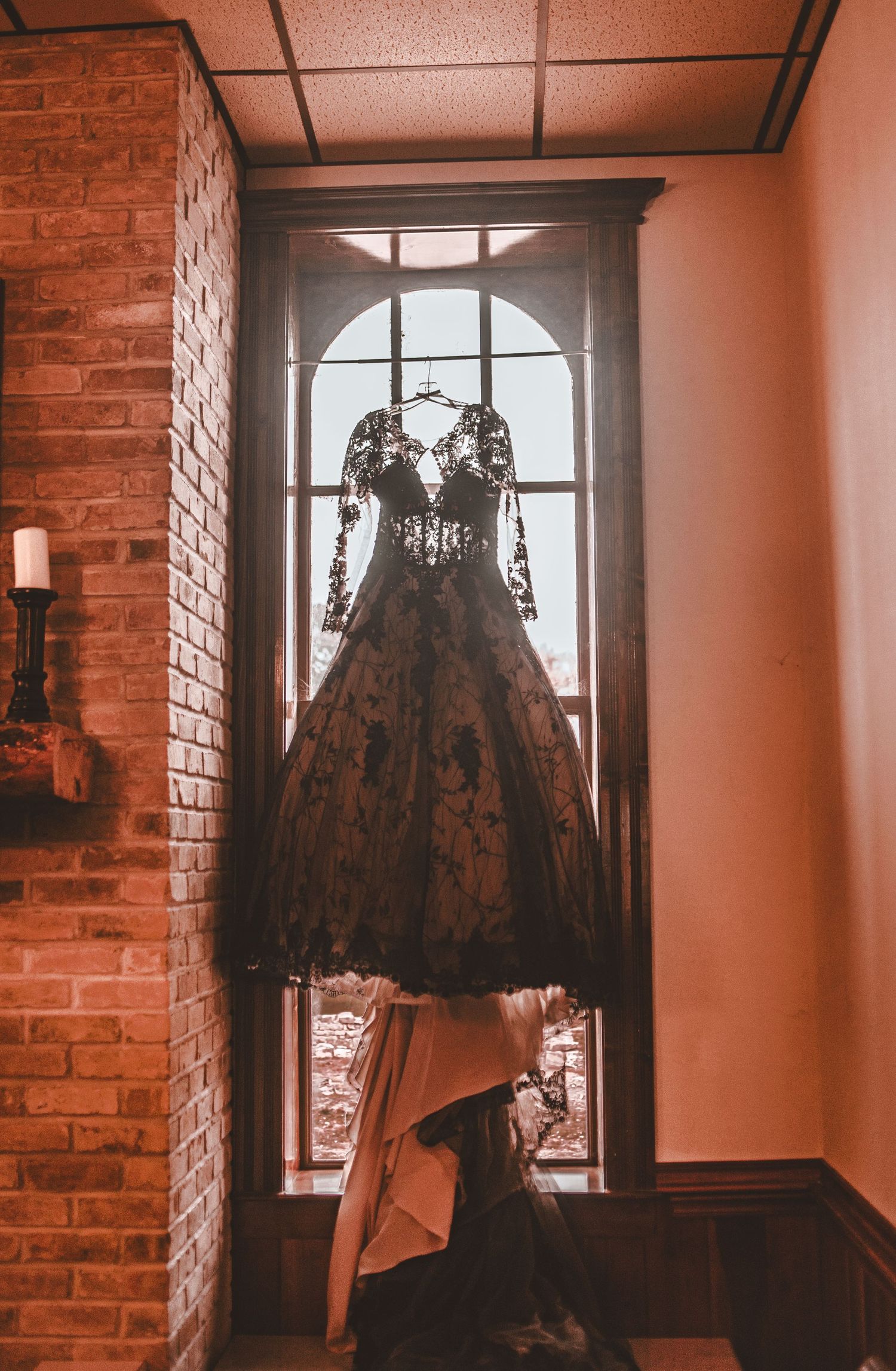 A vintage black lace dress hangs elegantly in front of an arched window against exposed brick walls.