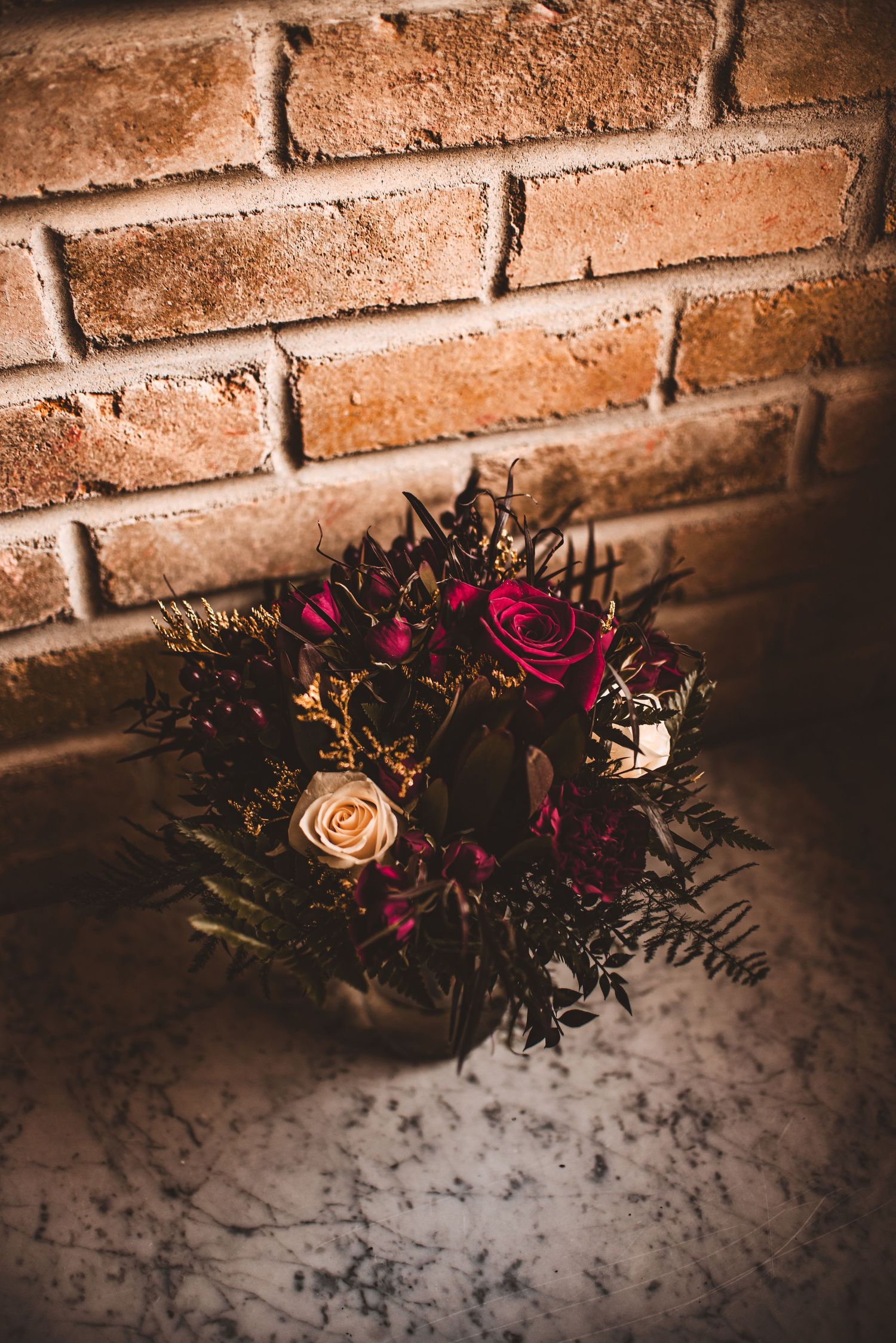 A moody floral arrangement with deep red roses against a rustic brick wall backdrop.