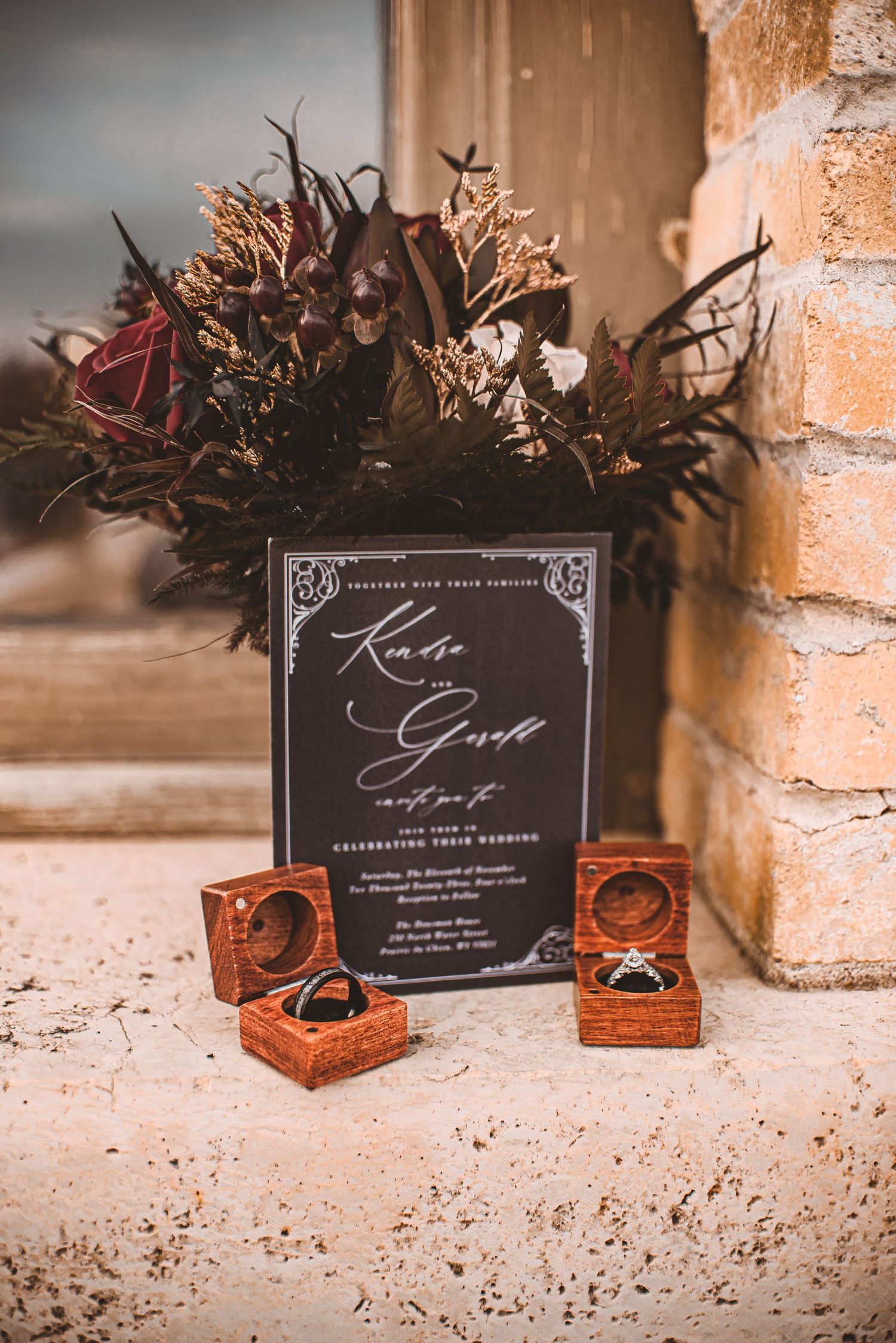 Wedding details with ring boxes, dried flowers, and a black invitation card against brick wall.