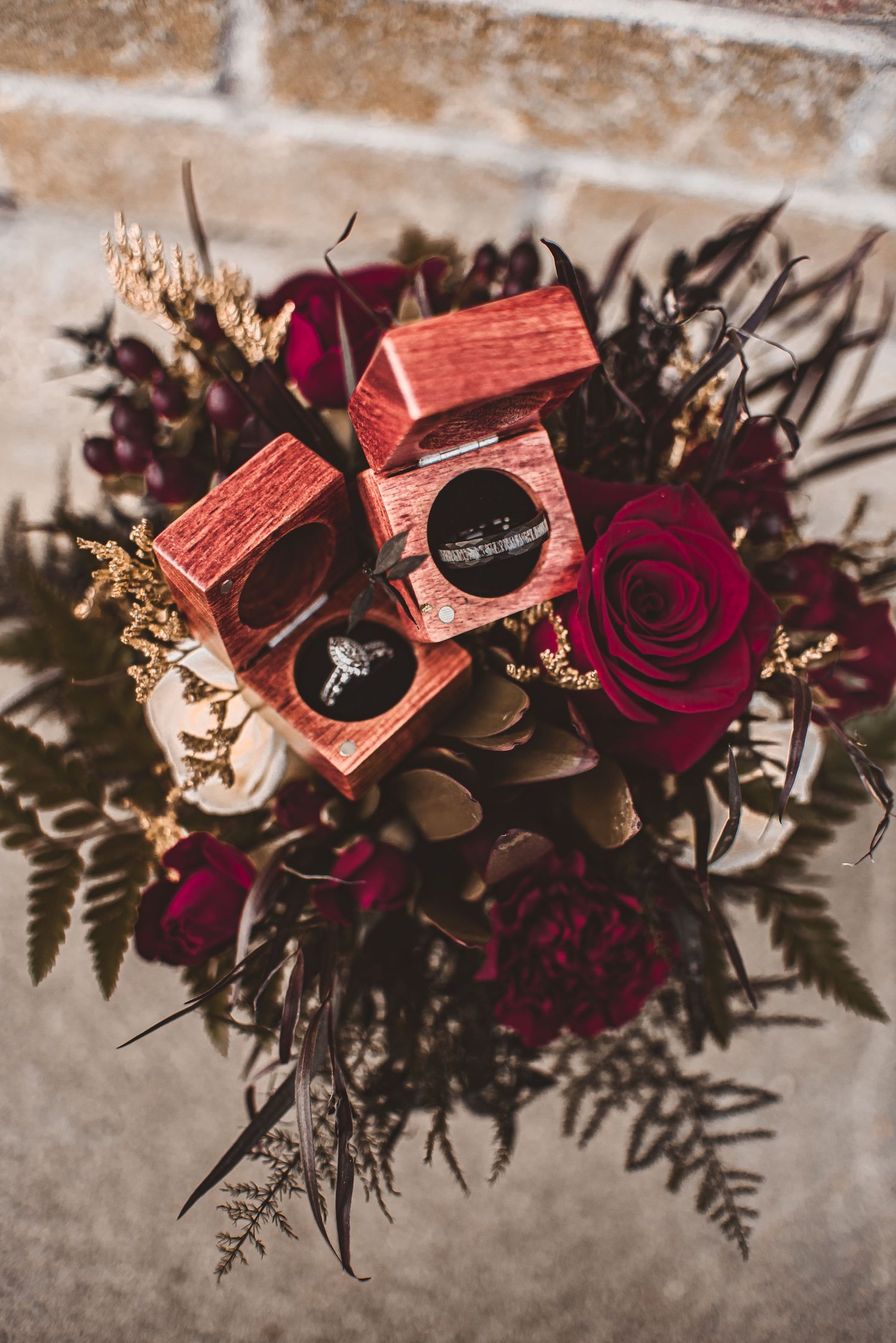 Wedding rings displayed in burgundy boxes amidst a dark romantic floral arrangement with red roses.
